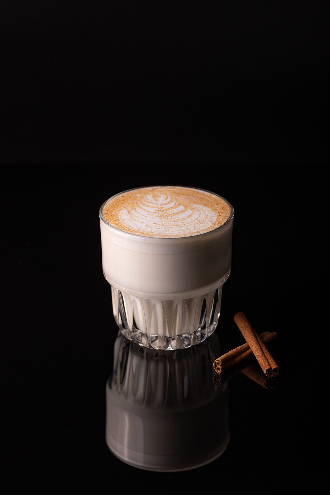 Cafea cu lapte, cu desen în spumă și scorțișoară presărată, lângă două batoane de scorțișoară