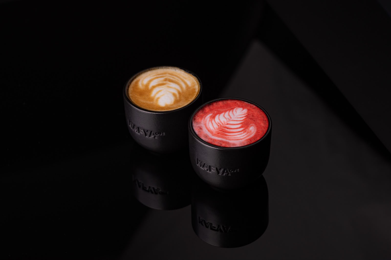 Două cești negre mate, gravate Kafya Cafe, cu latte art, reflectate pe o suprafață neagră lucioasă