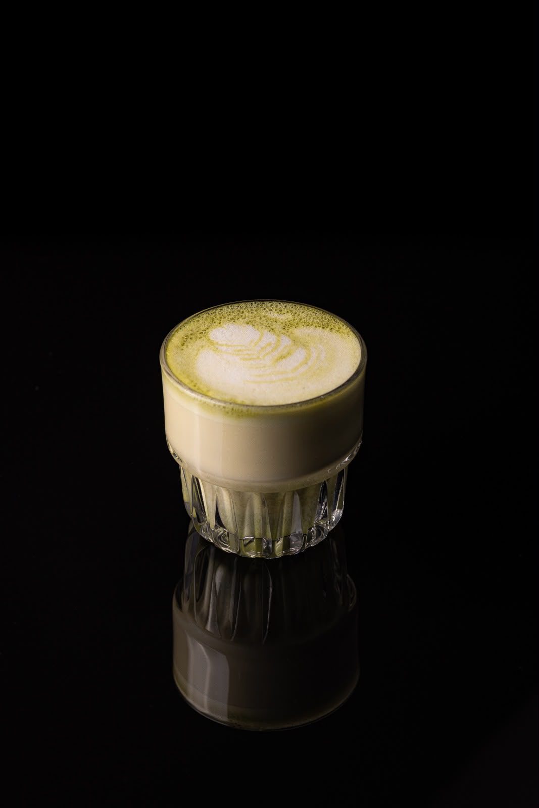 Matcha latte verde, cu desen în spumă, într-un pahar cu caneluri, pe fundal negru